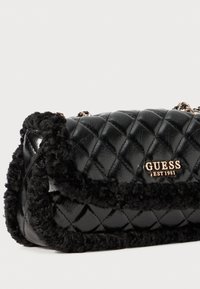 Borsa a tracolla nera trapuntata con rifiniture in peluche nero, hardware dorato e logo "GUESS EST 1981". Texture morbida con una finitura lucida. Forma rettangolare compatta.