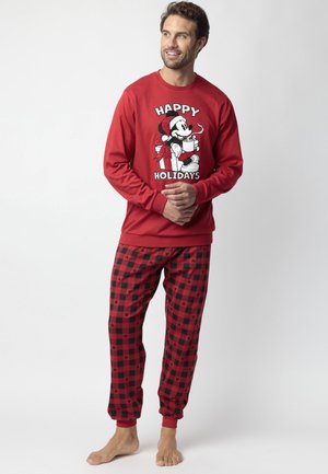 Uomo con felpa rossa da vacanza con grafica di Topolino e pantaloni del pigiama a quadri rosso-neri con piccoli icone di Topolino, in piedi a piedi nudi.