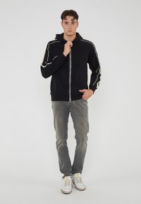 Felpa nera con zip e piping a contrasto color crema lungo le maniche, indossata sopra jeans skinny grigi e abbinata a sneakers bianche.