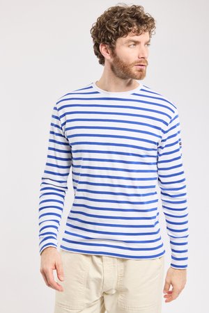 Uomo con capelli ricci che indossa una maglia bianca a maniche lunghe con strisce orizzontali blu e pantaloni beige chiaro, che guarda verso destra.
