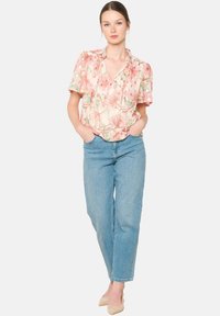 Kwitkowa bluzka w jasnoróżowym kolorze z falbaniastymi rękawami, zestawiona ze spodniami dżinsowymi w średnim odcieniu prostymi nogawkami i beżowymi szpilkami. Modelka stoi z dłońmi w kieszeniach.