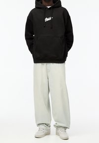 Osoba oblečená v černé mikině s bílým nápisem, světle šedých širokých džínách a bílých teniskách na čistém bílém pozadí.