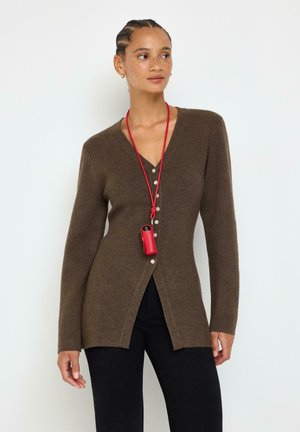 Donna con i capelli intrecciati indossa un cardigan marrone abbottonato e pantaloni neri, tenendo una cordicella rossa con una piccola custodia rossa intorno al collo.
