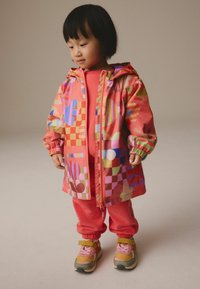 Bunter Windbreaker mit geometrischen Mustern in Rosa, Grün und Blau, kombiniert mit einem rosa Outfit und mehrfarbigen Sneakers mit rosa Schnürsenkeln.