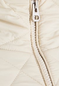 Beige quiltet jakke stof med diagonale syninger; har en sølvlynlås ned langs midten. Glat tekstur med en blød finish.