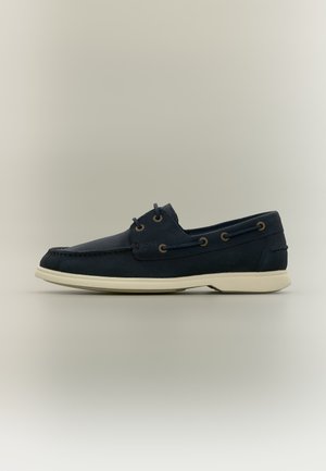 Navy blue leather boat shoe with white rubber sole, side lacing, and branded detail near the laces, shown in profile on a plain background.