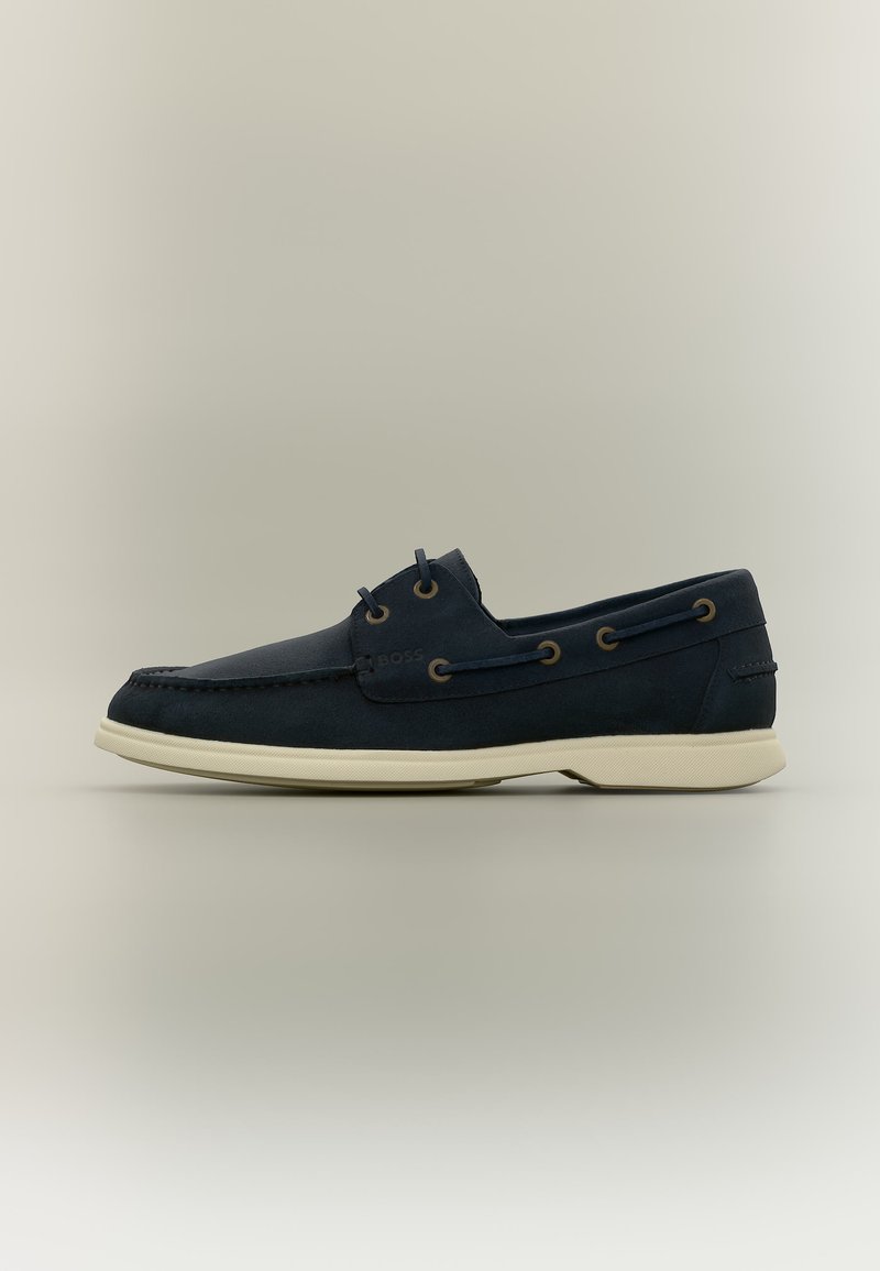 Chaussure bateau en cuir bleu marine avec semelle en caoutchouc blanche, laçage latéral et détail de marque proche des lacets, montrée de profil sur un fond uni.