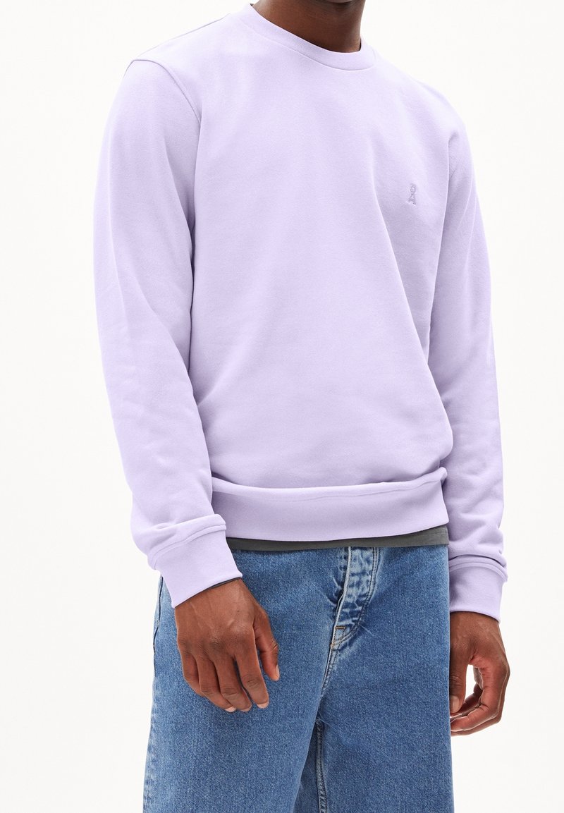 Ljus lavendelfärgad sweatshirt i mjukt tyg, med rund halsringning, ribbade ärmslut och fåll, samt en liten logga på bröstet, bärs med blå jeans.
