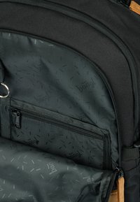 Interno di uno zaino nero con una texture liscia, una tasca con zip e foderatura con motivo caratterizzato da piccoli loghi neri sparsi.
