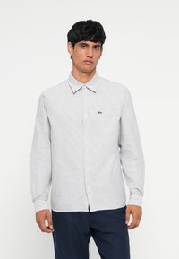 Striped long-sleeve shirt in white and navy, featuring a button-down collar, front buttons, and a small green logo on the chest pocket.