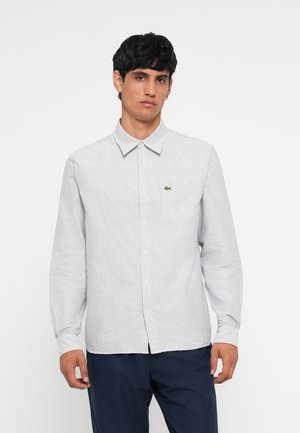 Camisa de manga larga a rayas en blanco y azul marino, con cuello abotonado, botones en el frente y un pequeño logo verde en el bolsillo del pecho.