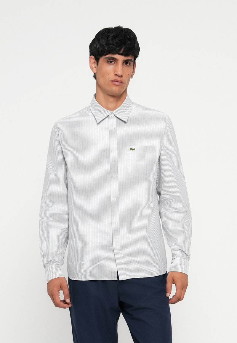 Striped long-sleeve shirt in white and navy, featuring a button-down collar, front buttons, and a small green logo on the chest pocket.