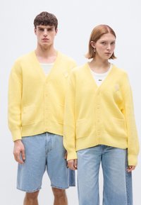 Κίτρινα πλεκτά ζακέτες με ραβδώσεις και V-λαιμόκοψη, με τσέπες στο μπροστινό μέρος, κουμπιά και μικρό λογότυπο. Φοριούνται πάνω από ανοιχτόχρωμα τοπ.