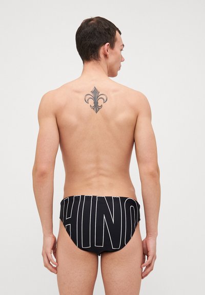 Boxers de bain noirs avec des lettres blanches à l'arrière. Le matériau est lisse et le design est ajusté avec une coupe simple.
