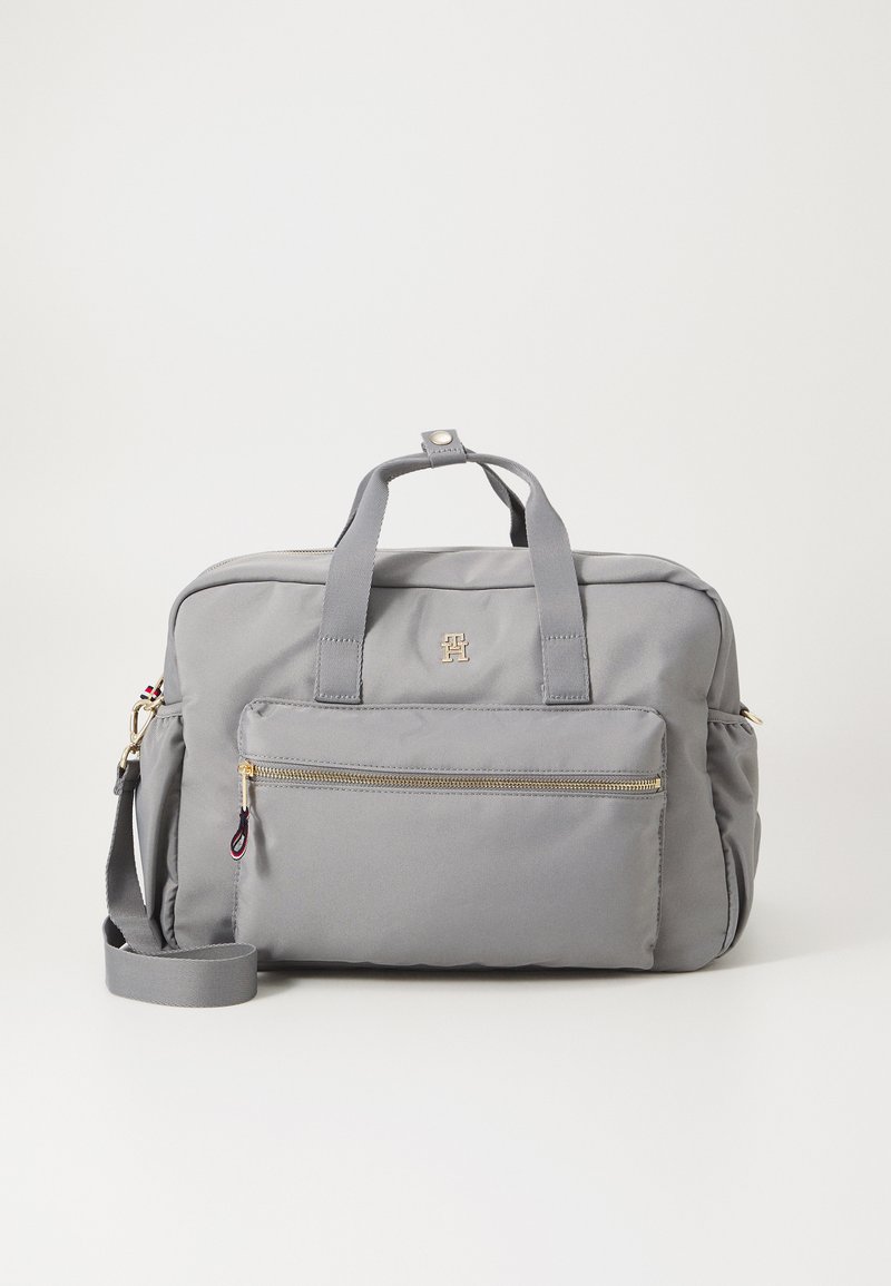 Grå nylon taske med dobbelthåndtag, en aftagelig strop, en frontlomme med lynlås og hardware i guldfarve. Glat tekstur med rene linjer.