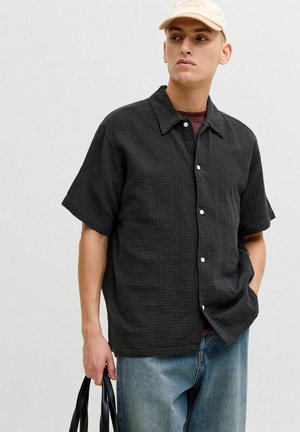 Homme portant une casquette beige, une chemise noire ample à manches courtes et boutonnée, un jean bleu, tenant un sac fourre-tout noir d'une main.