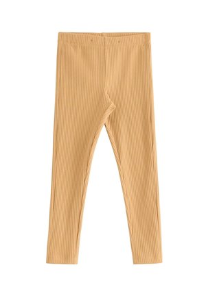 Ung pige med langt hår i fletninger iført en hvid blomsterstrik, sennepsfarvede ribstrikkede leggings og mørkerøde sneakers, stående mod en ensfarvet baggrund.