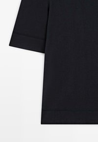 Zwart katoenen T-shirt met korte mouwen, met zichtbare stiksels op de linker mouw en onderzoom, op een witte achtergrond.