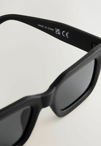 Lunettes de soleil noires avec une finition mate, verres rectangulaires et branches larges et robustes. "FABRIQUÉ EN CHINE UK CA CE" gravé sur l'intérieur de la branche.