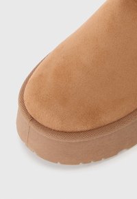 Botte en suède marron clair avec un bout rond, un dessus texturé et une semelle en caoutchouc plate, présentant un design simple et épuré avec des accents cousus.
