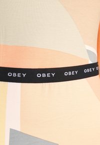 Grafika materiału z jasnymi pastelowymi kolorami—brzoskwiniowym, miętowym i szarym—wykończona czarną gumką, na której widnieje napis "OBEY" w białym tekscie.