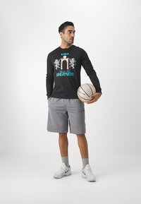 Black long-sleeve shirt with graphic design and "Living for Greatness" text, paired with gray shorts and white athletic shoes. Holding a basketball.
