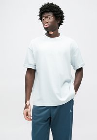 Ανοιχτόγαλάζιο βαμβακερό μπλουζάκι με γυρισμένα μανίκια, με μικρό λογότυπο Jordan στο στήθος. Συνδυάζεται με σκούρο μπλε αθλητικό παντελόνι.