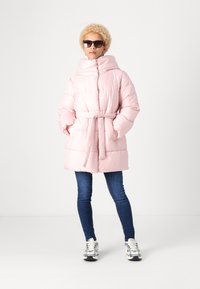 Giacca imbottita e con cintura di colore rosa pallido, con colletto alto e texture liscia. Indossata con jeans blu e sneakers bianche. Occhiali da sole inclusi.