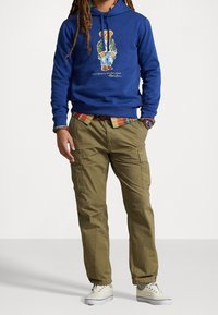 Person som bär en blå hoodie med björnmotiv, rutig skjorta under, olivfärgade cargobyxor, vita sneakers och en armbandsklocka, stående mot vit bakgrund.