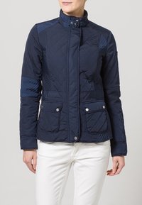 Granatowa pikowana kurtka z stojącym kołnierzem, dwoma przednimi kieszeniami i kontrastowymi panelami. Posiada zapięcia na zatrzaski oraz miękką fakturę.