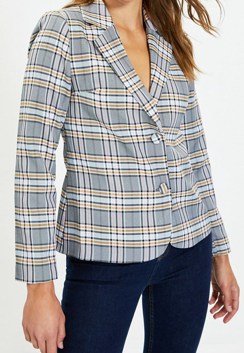 Femme portant un blazer à carreaux gris, blanc et moutarde avec deux boutons, associé à un jean bleu foncé.