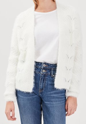 Woman wearing a white fuzzy knit cardigan over a plain white T-shirt paired with high-waisted blue jeans with double buttons.