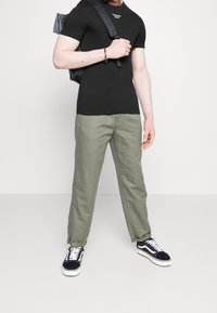 T-shirt noir ajusté avec logo blanc, pantalon décontracté vert olive, et baskets noires et blanches. Sac à dos sur l'épaule, mains jointes.