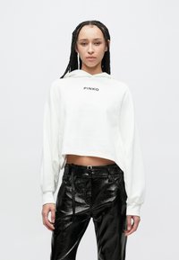 Balta īsā kapuce ar melnu "PINKO" logotipu, kombinēta ar spīdīgām melnām augsta vidukļa mākslīgās ādas biksēm, kurās ir priekšējās kabatas un gluda tekstūra.