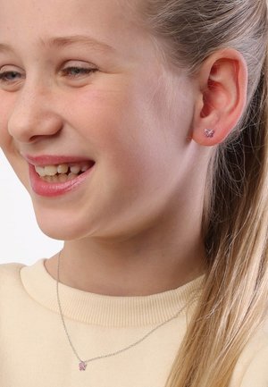 Vlinder oorbellen in roze edelstenen bij een meisje, gecombineerd met een zilveren ketting met een bijpassende hanger. Lichtgele trui.
