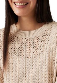 Persona sorridente che indossa un maglione lavorato a maglia beige con scollatura a costine e intricati motivi verticali simili a pizzi.