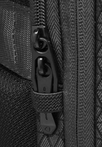 Fermeture éclair noire sur un tissu texturé, avec un curseur en plastique portant un petit logo. Gros plan sur les dents de la fermeture éclair et les détails de la couture.