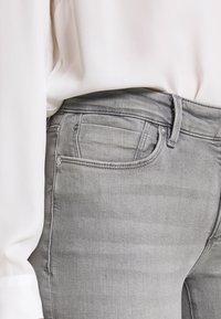 Jeans de mezclilla gris con un diseño ajustado, que presentan costuras sutiles, un bolsillo para monedas y una textura suave. Combinados con una blusa blanca.