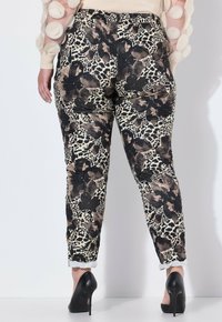 En kvinna som bär leopard- och blommönstrade byxor tillsammans med svarta högklackade skor, står mot en vit bakgrund.