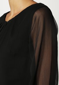 Gros plan d'un chemisier noir avec manches longues semi-transparentes et encolure ronde sur le haut du torse et l'épaule d'une personne.