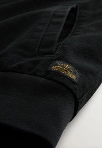 Černá strukturovaná tkanina má prošívaný lem a obdélníkovou tkanou nálepku se zlatým textem "ESTD 1992 STANDARD SUPPLY CO."