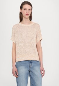 Lys beige strikket kortermet genser med løs passform og ribbet halskant, parret med lys blå denim. Teksturert stoff med åpen veving.