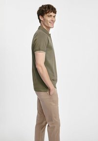 Oliwkowa koszulka polo z kontrastowymi jasnymi wykończeniami, połączona z jasnobrązowymi spodniami. Materiał wydaje się być miękki, z klasycznym krojem i krótkimi rękawami.
