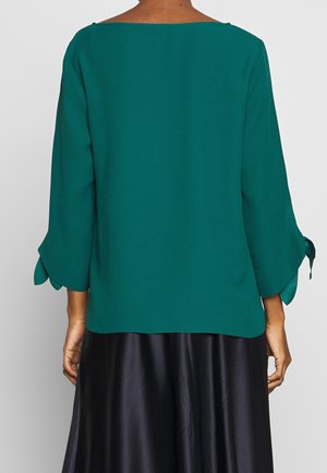 Achteraanzicht van een persoon die een tealblauwe blouse met lange mouwen draagt, voorzien van gestrikte mouwdetails, en een zwarte satijnen rok.