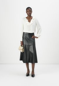 Cremfarbene Bluse mit langen Ärmeln, Rüschendetails und Schleife, kombiniert mit einem hoch taillierten schwarzen Leder-A-Linienrock und einer beigen Handtasche.