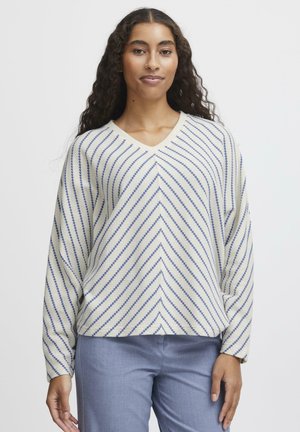 Maglione a V in crema con strisce diagonali blu. Tessuto morbido e tessurizzato con maniche lunghe, abbinato a pantaloni azzurri chiari.