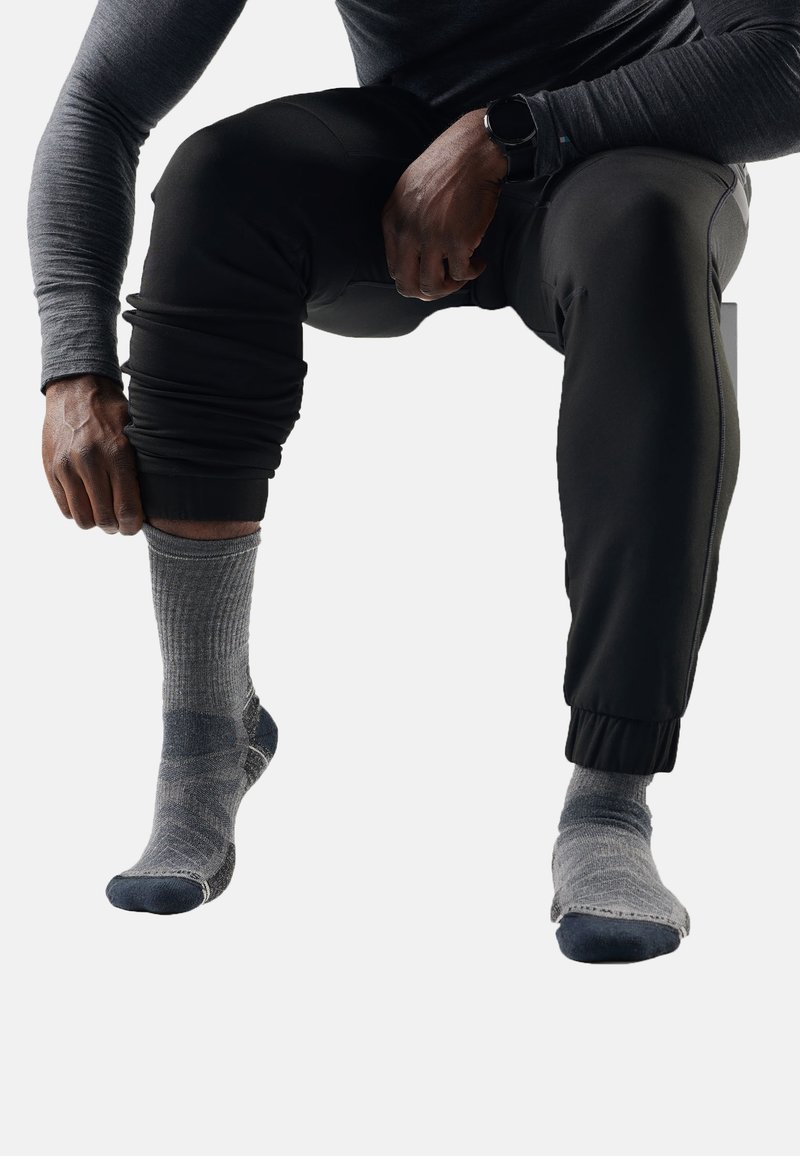 Persona che indossa pantaloni neri e calzini grigi sistema il calzino sul piede sinistro mentre è seduta davanti a uno sfondo neutro.