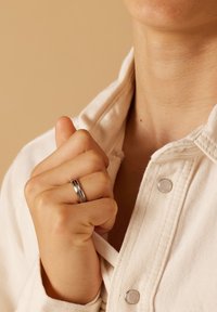 Zilveren ring met een gladde afwerking en twee horizontale groeven, gedragen op een gebalde hand, tegen een neutrale beige achtergrond.