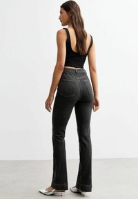 Jean évasé en denim à lavage sombre avec une taille haute, un tissu texturé et deux poches arrière. Associé à un top sans manches noir et des chaussures argentées.