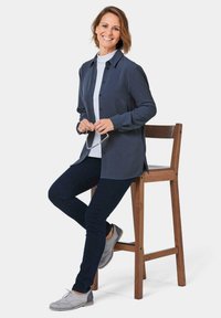 Dunkelblaues Hemd mit Knöpfen über einem weißen Rollkragenpullover, kombiniert mit dunklen Jeans und hellgrauen Schuhen, sitzt auf einem Holzbarhocker.